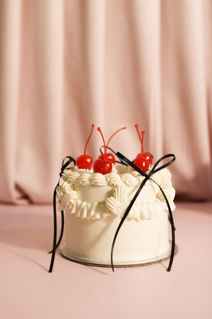 White cake with white cream ruffles, three black ribbons, and five red cherries on top of the cake